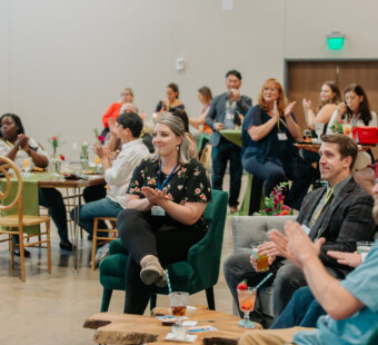 People sitting and standing in a room, clapping and watching an event, drinks on tables, casual setting.