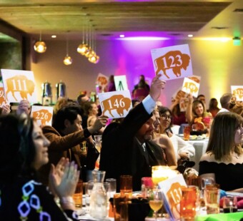 People at an auction hold up numbered signs while seated at brightly lit tables in a lively event space.