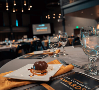 Table set with dessert, water glasses, and an awards program at a formal event, with blurred screens in background.
