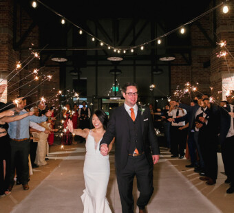 A bride and groom walk hand in hand through cheering guests holding sparklers at night.