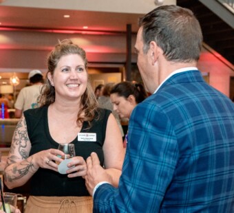 A woman smiling and talking to a man at a social event in an indoor setting.