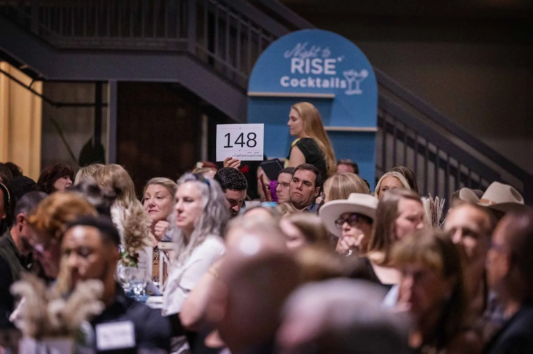 A blue cocktail wall sits in the background while a group of people sit at tables, and one holds up a paddle for a live auction at a nonprofit Gala